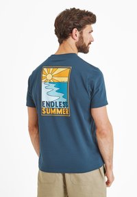 Man wearing navy blue t-shirt with "Endless Summer" sun and ocean graphic on back, paired with beige shorts, standing in profile.