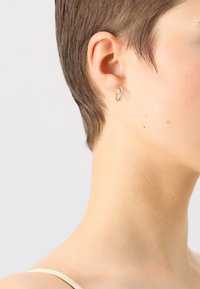 Goldene Creolen mit strukturierter Oberfläche, die kleine, funkelnde Akzente auf der Vorderseite aufweisen. Getragen von einem Model mit kurzem Haar.