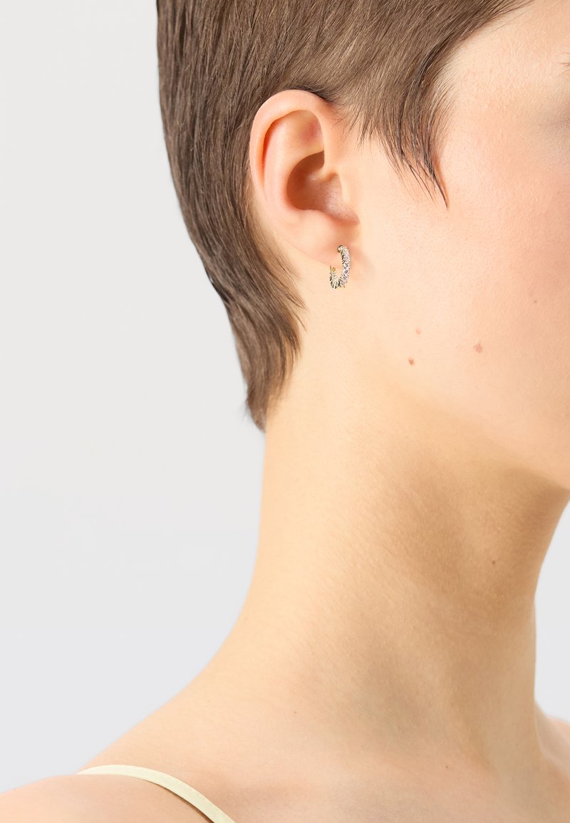 Goldene Creolen mit strukturierter Oberfläche, die kleine, funkelnde Akzente auf der Vorderseite aufweisen. Getragen von einem Model mit kurzem Haar.