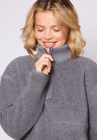 Pullover grigio in pile con colletto alto, dotato di chiusura a mezza cerniera. Il tessuto ha una finitura morbida e testurizzata con dettagli a coste.