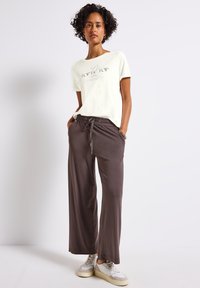 Lys cremefarvet t-shirt med sort print, parret med løse brune sweatpants med elastisk talje og snor. Hvide sneakers med beige detaljer.