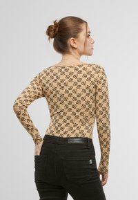 T-shirt beige à manches longues avec des logos noirs dans un motif répétitif. Design ajusté avec un col rond, assorti à un pantalon noir.