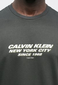 Ciemnoszary bawełniany t-shirt z okrągłym dekoltem, z wyraźnym białym i jasnooliwkowym napisem: "CALVIN KLEIN NEW YORK CITY SINCE 1968."
