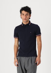 Polo de color azul marino hecho de algodón, con una tapeta de tres botones, cuello y mangas a rayas rojas y blancas, y un pequeño detalle del logo.