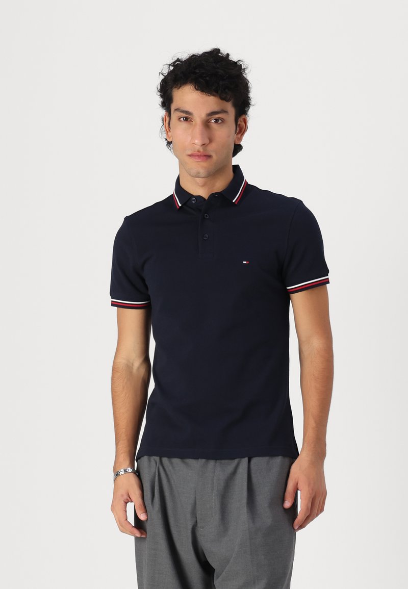 Polo de color azul marino hecho de algodón, con una tapeta de tres botones, cuello y mangas a rayas rojas y blancas, y un pequeño detalle del logo.