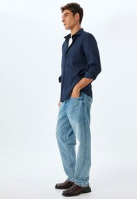 Camisa de botones azul marino, pantalones vaqueros rectos de un azul claro y zapatos de cuero marrón. La camisa tiene un cuello clásico; los vaqueros presentan un desgaste sutil.