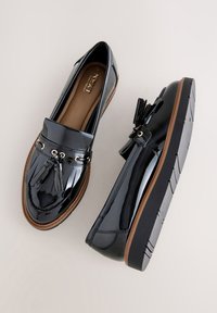 Mocassins en cuir verni noir avec pompons, bout rond et accents de matériel doré ; semelle texturée avec bordure marron. Design plat.