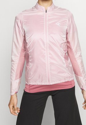 Personne portant une veste zippée rose clair sur une chemise rose plus foncée et un pantalon noir, debout devant un fond uni.