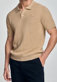Béžové žebrované polo tričko s klasickým límcem, krátkými rukávy a logem na hrudi; látka vypadá texturovaně a přiléhavě.