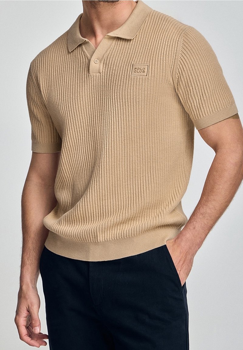 Béžové žebrované polo tričko s klasickým límcem, krátkými rukávy a logem na hrudi; látka vypadá texturovaně a přiléhavě.