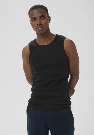 Homme aux cheveux courts portant un débardeur noir sans manches et un pantalon bleu foncé, debout les mains derrière le dos devant un fond uni.