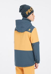 Farbbesetzte Jacke in Gelb und Blau mit Kapuze, ausgestattet mit elastischen Bündchen. Eine blaue Strickmütze mit "PRO" in orangefarbener Schrift.