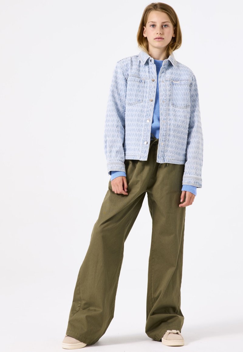 Lichtblauwe spijkerjack met een gestructureerd patroon, knoopsluiting, twee borstzakken; gecombineerd met oversized groene broek en beige schoenen.