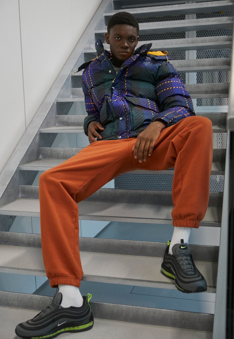 Padded jacket w niebiesko-zielonym geometrycznym wzorze, pomarańczowe dresy i czarne sneakersy z zielonymi akcentami, siedzący na metalowych schodach.