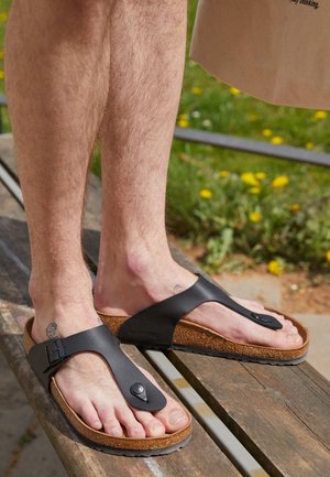 Bare male feet wearing black cork-soled thong sandals resting on a wooden bench outdoors with green grass and yellow flowers in the background.