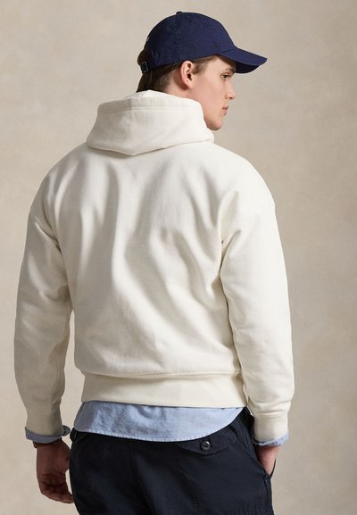 Κρεμ φούτερ με ριμπ τελείωμα και μανσέτες, με κουκούλα και κορδόνι. Φορεμένο πάνω από ανοιχτό μπλε πουκάμισο, με σκούρο παντελόνι. Στο κεφάλι μπλε σκουφάκι.