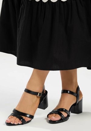 Stopy w czarnych, odkrytych sandałkach na obcasie z paskami wokół kostek, pod czarną spódnicą z falowanym białym brzegiem.