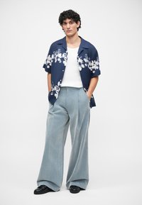 Chemise à motifs floraux navy à manches courtes, débardeur blanc, pantalons larges bleu clair, et chaussures noires. Tissu lisse et coupe décontractée.