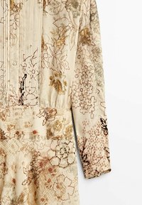 Robe longue à manches longues de couleur crème avec devant boutonné et imprimé floral dans des tons de marron, beige et orange doux.