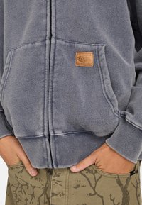 Grå zip-up hættetrøje med tekstureret stof og et syet logo lap på brystet, båret over khaki bukser med en grenmønster.