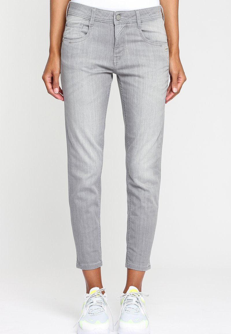 Femme portant un jean slim gris clair et des baskets blanches avec des accents jaune néon, debout devant un fond uni.