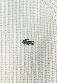 Weißer gestrickter Pulloverstoff mit gerippter Textur. Verfügt über ein kleines grünes gesticktes Krokodil-Logo mit roten Akzenten.