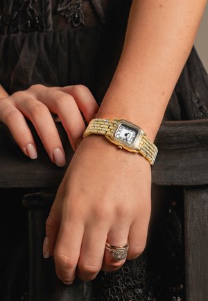 Montre en or avec un cadran carré, un cadran blanc et une lunette embellie. Le bracelet en métal présente un design texturé et de petits accents scintillants.