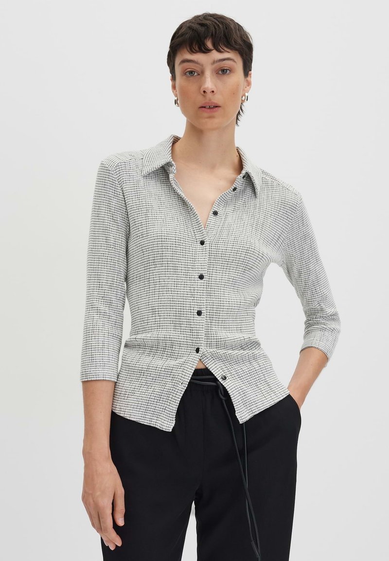 Camicia aderente con bottoni in un motivo a griglia bianca e nera. Presenta un colletto a punta e maniche a tre quarti, realizzata in un tessuto testurizzato.