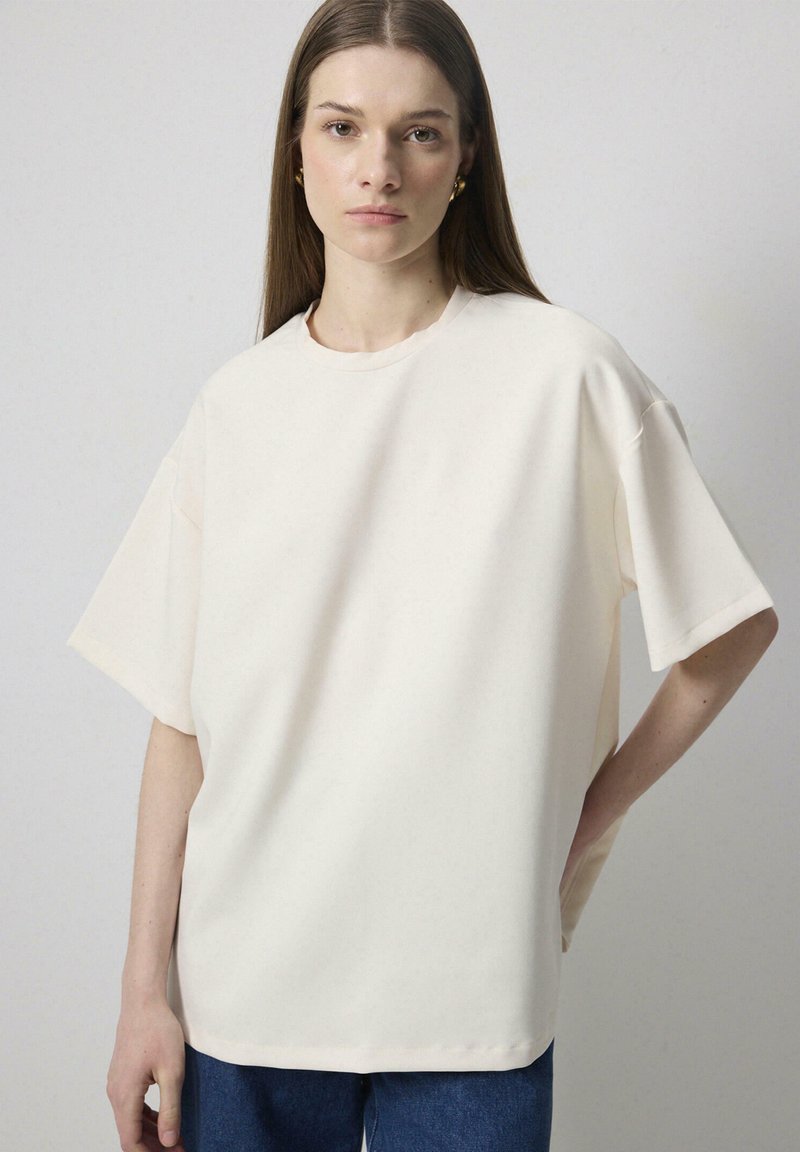 Γυναίκα με μακριά καστανά μαλλιά που φοράει απλό φαρδύ εκρού T-shirt και μπλε τζιν, μπροστά σε ουδέτερο φόντο.