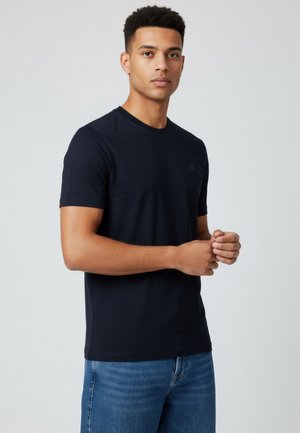 Joven con cabello corto y rizado, vestido con una camiseta negra lisa y vaqueros azules, de pie frente a un fondo gris claro.