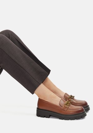 Brązowe skórzane mokasyny z masywną czarną podeszwą, ozdobione złotym łańcuszkiem na cholewce. Połączone z eleganckimi ciemnymi spodniami.