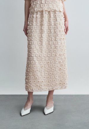 Világos rózsaszín midi szoknya háromdimenziós virágos textúrával, fehér hegyes orrú cipőkkel párosítva, csipke díszítéssel.