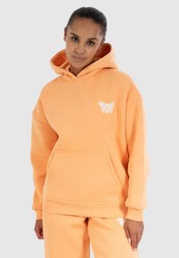 Oranje hoodie van zacht materiaal, met een voorzak en een wit bloemmotief op de borst. Verstelbare trekkoordcapuchon.
