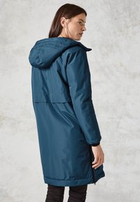 Teal vattentät parka med huva, med en midjeresår, långa ärmar och två sidofickor med dragkedja. Slät textur och knä-lång design.