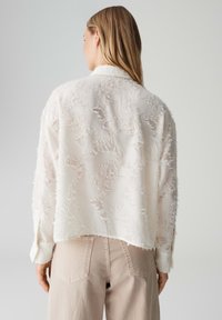 Blusa bianca testurizzata con vestibilità ampia e bordi sfrangiati, dotata di colletto e maniche lunghe, abbinata a pantaloni beige chiaro a vita alta.
