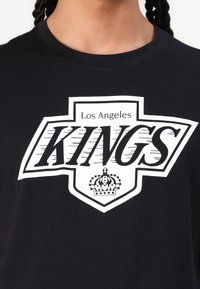 Crna pamučna majica s velikim bijelim grafičkim prikazom koji sadrži tekst "Kings" i "Los Angeles", u obliku štita s detaljem krune na dnu.
