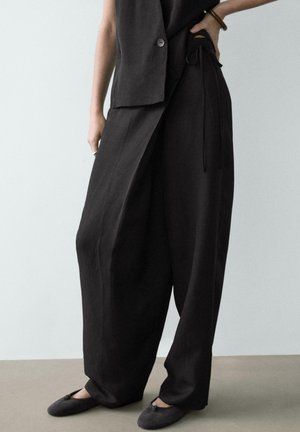 Person trägt lockere schwarze weite Hosen, passende schwarze ärmellose Weste und schwarze flache Schuhe, steht mit einer Hand in der Hüfte.