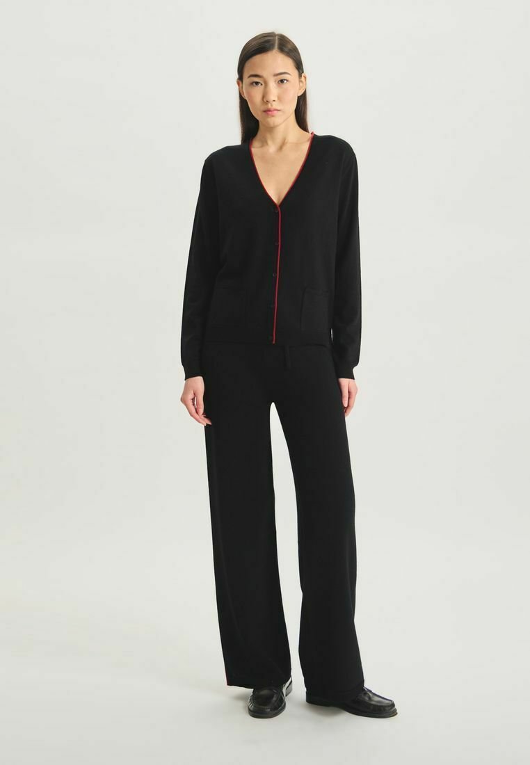Cardigan noir avec des bordures rouges, décolleté en V, manches longues et poches avant associé à un pantalon large noir et des chaussures noires.
