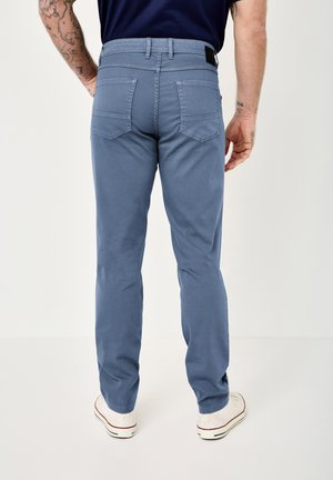 Man wearing light blue slim-fit pants and white sneakers, standing facing away with visible tattoos on arms and dark blue shirt tucked in.