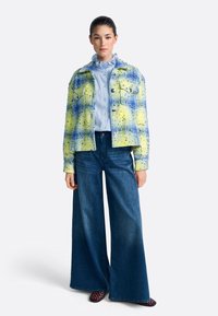 Barven tweed suknjič v rumeni in modri barvi, z ohiščno rezjo in dvema žepoma na prsih, v kombinaciji s svetlo modro srajco z volančki in širokimi kavbojkami.