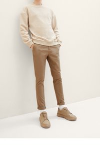 Osoba v béžovém svetru, hnědých nohavicích na ohrnutí, bílých ponožkách a béžových teniskách, stojící proti krémové zdi.