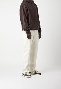 Persona vistiendo una sudadera marrón, pantalones sueltos color blanco roto y zapatillas bicolores en negro, marrón y blanco, de pie contra un fondo blanco liso.