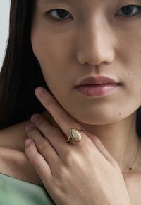 Zlatý prsteň s textúrovanou kruhovou plochou a stredovým kameňom, nosený na ruke, ktorá spočíva blízko tváre, na tlmenom pozadí.