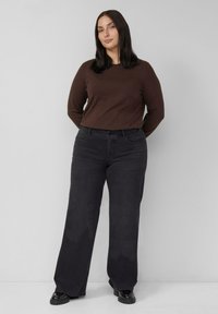 Brązowa bluzka z długim rękawem zestawiona z luźnymi ciemnoszarymi dżinsami. Strój cechuje się prostym krojem i miękką fakturą tkaniny. Całość dopełniają czarne buty.