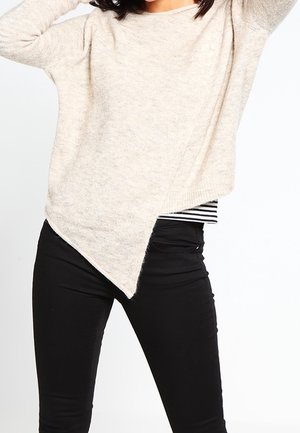 Vrouw die een beige asymmetrische trui draagt over een zwart-wit gestreept shirt en een zwarte broek, poserend met opgeheven armen.