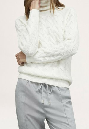 Personne portant un pull à col roulé blanc en maille torsadée et un pantalon gris clair à cordon, les mains partiellement croisées.