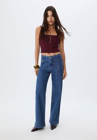 Top a busto di un rosso scuro con spalline sottili, abbinato a jeans in denim blu a vita alta con gambe larghe, dotati di due tasche frontali e dettagli in oro.