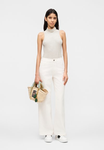 Γυναίκα με αμάνικη ριγέ ζιβάγκο, λευκό παντελόνι φαρδιάς γραμμής, λευκά αθλητικά παπούτσια, κρατώντας πλεκτή τσάντα με πολύχρωμες λαβές.