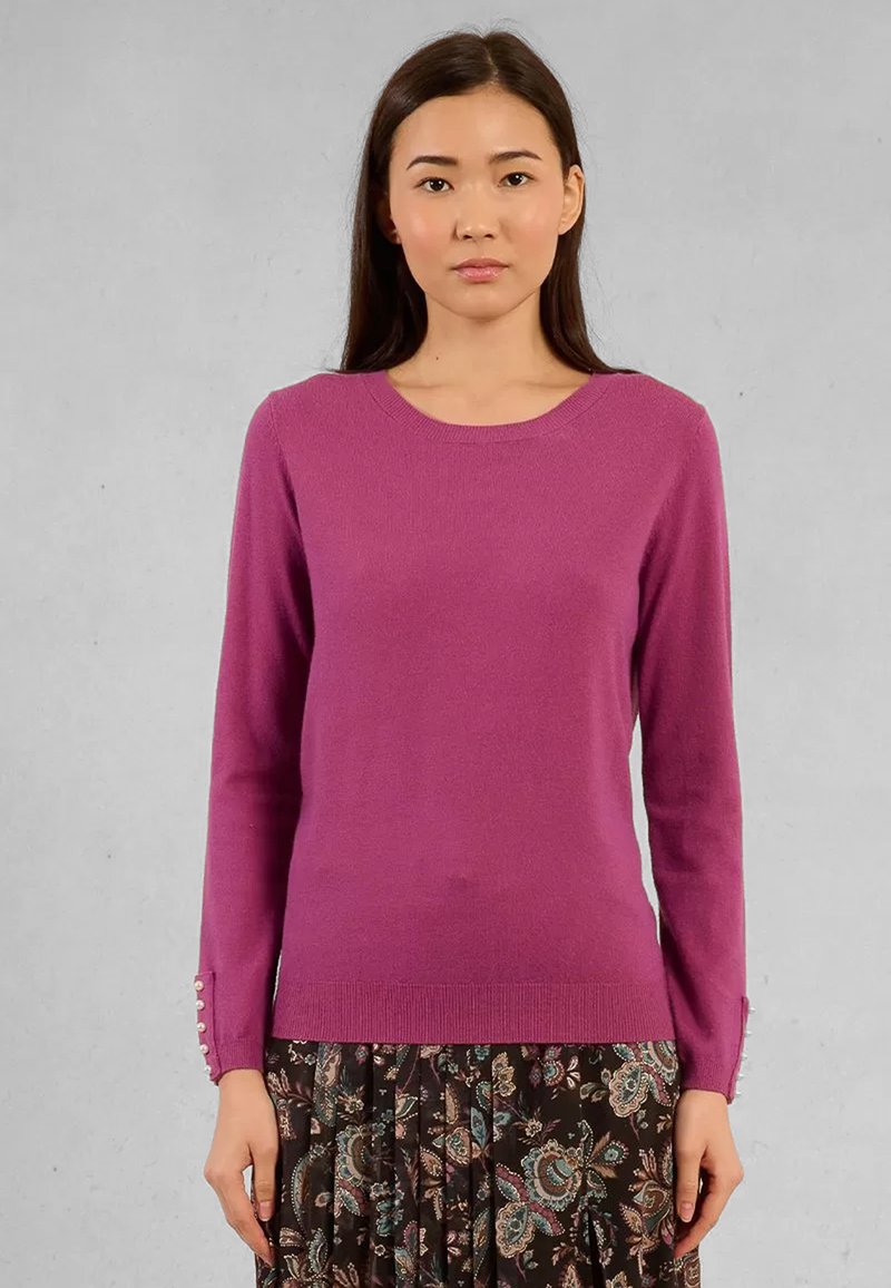 Donna che indossa un maglione viola a maniche lunghe con bottoni perlati sui polsini e una gonna a fantasia floreale scura su uno sfondo grigio semplice.