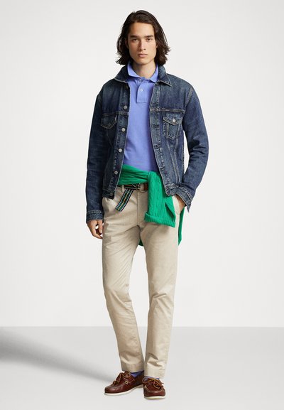 Kurtka dżinsowa, jasnoniebieska polo, beżowe spodnie, zielony sweter zawiązany wokół talii, brązowe buty żeglarskie. Codzienny strój z warstwową stylizacją.